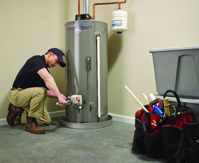Alt Text: A man in a cap and work clothes kneels to adjust the controls on a tall, gray gas water heater in a utility space, with a toolbox nearby.