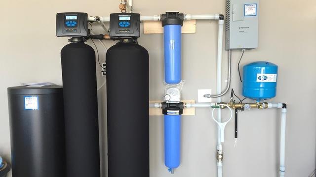 A robust residential water treatment system featuring two tall black tanks, two blue filter housings, and a blue pressure tank, neatly installed against a light-colored wall.