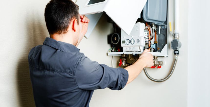 A person in a dark shirt is seen from behind, looking into and working on the open white cover of a modern wall-mounted boiler or water heater, revealing its internal components, wires, and pipes.