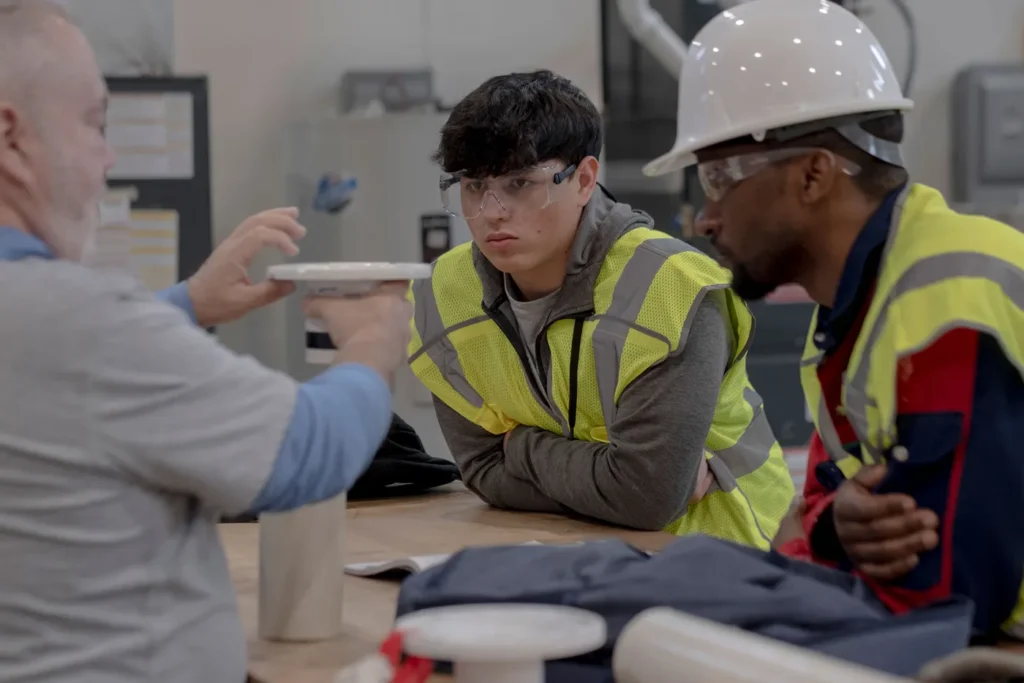 Three men, two wearing safety gear, appear to be in a training or discussion session related to construction or a trade.
