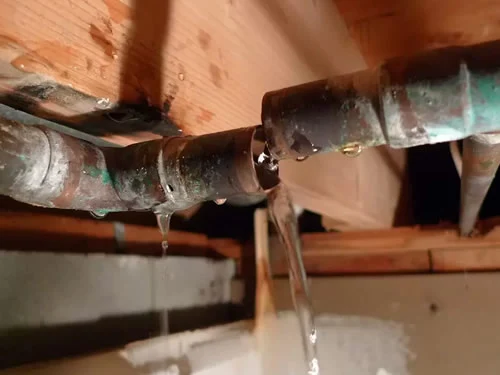 A close-up of a burst and leaking copper pipe in a basement or crawl space, with water actively gushing and dripping onto the surrounding area.