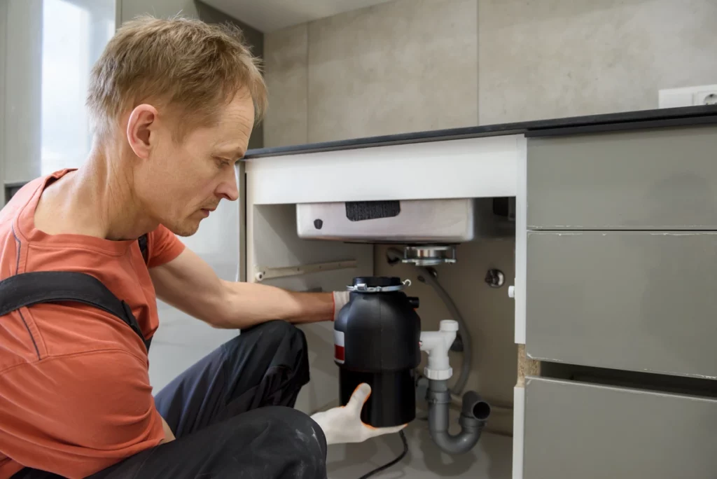A man in an orange shirt and black overalls is installing or servicing a black garbage disposal unit under a modern kitchen sink.