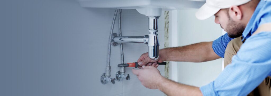 A plumber wearing a white cap and blue shirt is working on the chrome P-trap and water supply lines beneath a white sink, using a wrench to tighten a fitting.