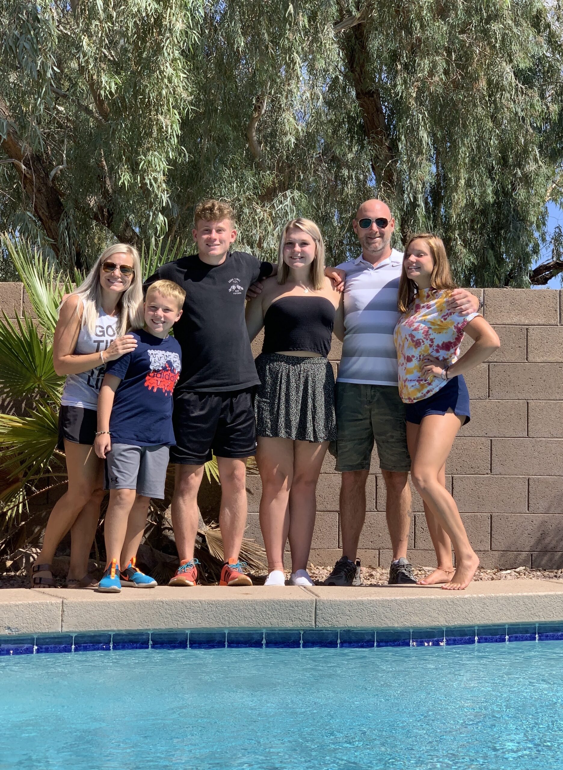 A family of six, consisting of two adults and four children/teenagers, stands smiling by the edge of a swimming pool on a sunny day.
