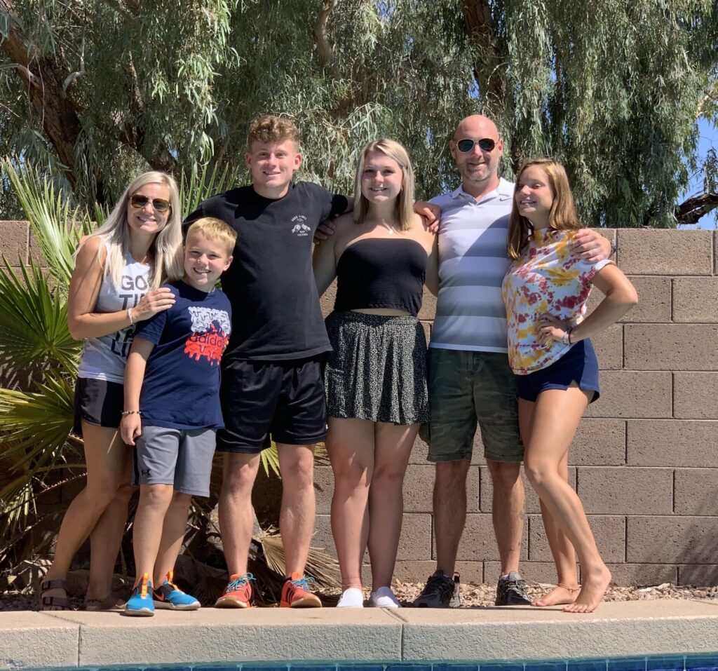 A family of six, two adults and four children/teenagers, stands smiling by a swimming pool with a block wall and trees in the background.