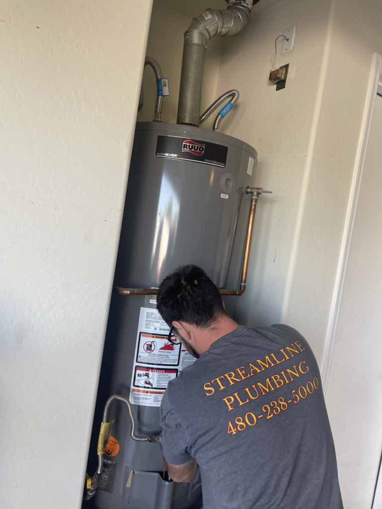 A plumber wearing a "Streamline Plumbing" t-shirt works on installing a new Ruud water heater in a narrow utility closet.
