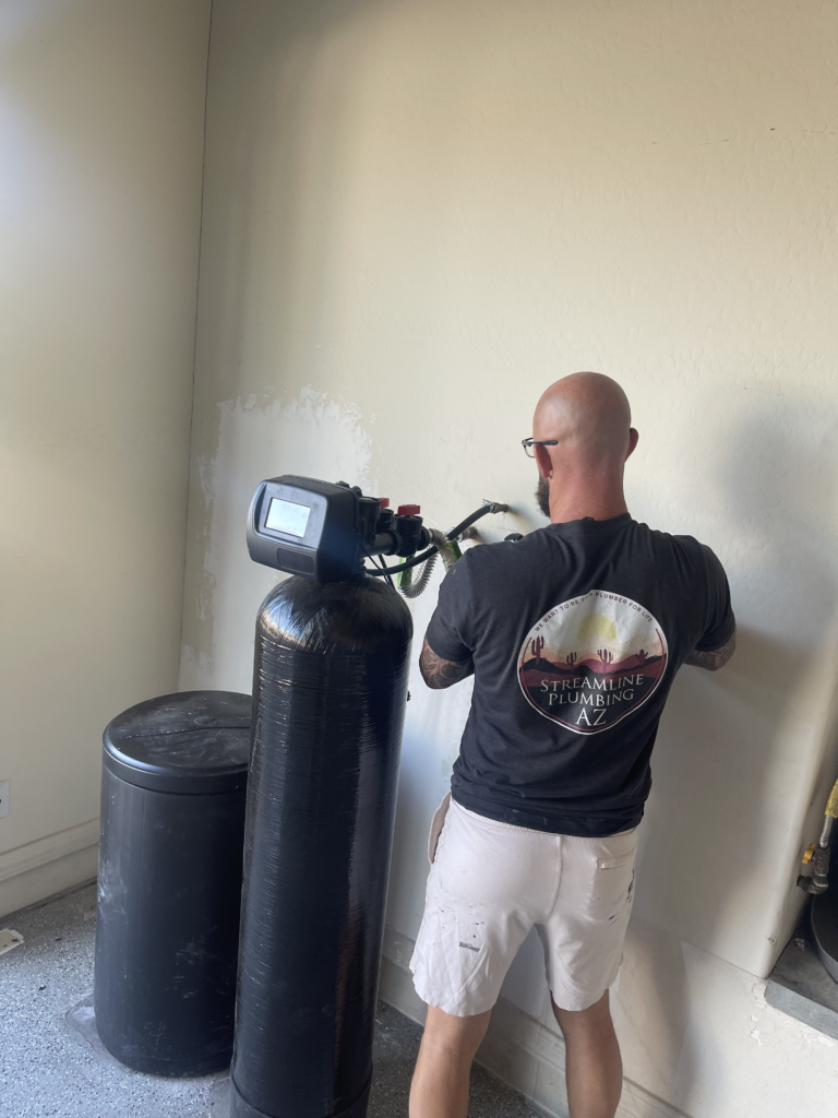 A bald man in a "Streamline Plumbing" t-shirt works on a wall next to a large black water softener system.