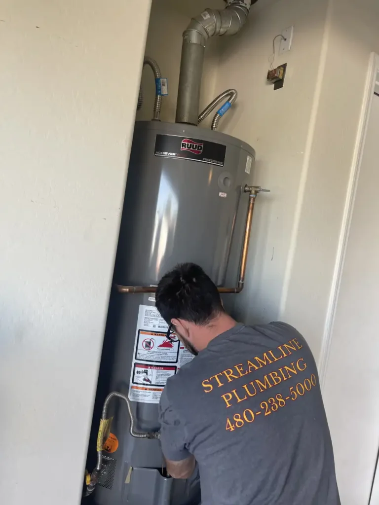 A plumber wearing a "Streamline Plumbing" t-shirt works on installing a new Ruud water heater in a narrow utility closet.