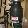 A close-up view under a kitchen sink showing a newly installed or recently serviced dark gray garbage disposal unit with a red "Call for Service" sticker for Apex Plumbing & Sewer Inc.