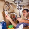 A man with dark hair looks up intently while wiping a section of a chrome P-trap pipe under a sink with a white paper towel, checking for leaks.