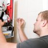 A male technician with a screwdriver works on the internal components of a wall-mounted gas boiler or central heating unit.