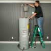 A man on a green step ladder is working on the copper pipes and expansion tank above a gray gas water heater.