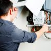 A person in a dark shirt is seen from behind, looking into and working on the open white cover of a modern wall-mounted boiler or water heater, revealing its internal components, wires, and pipes.