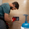 A male technician in work attire and red gloves is installing or adjusting a black flush plate or control panel on a light wood-paneled wall, likely in a modern bathroom.