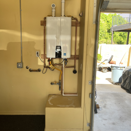 A white, rectangular tankless water heater is mounted on a yellow wall in a garage, with copper and PVC pipes connecting to it, and an open garage door reveals an outdoor view.