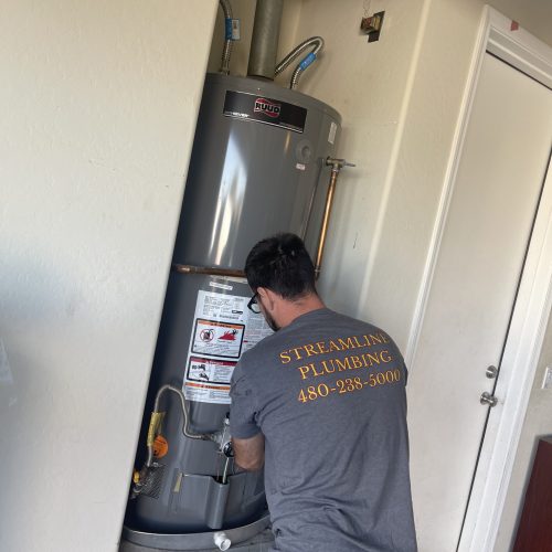 A plumber, wearing a "Streamline Plumbing" t-shirt, kneels to work on the lower section of a gray Ruud water heater in a narrow utility space.