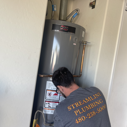 A plumber wearing a "Streamline Plumbing" t-shirt works on installing a new Ruud water heater in a narrow utility closet.