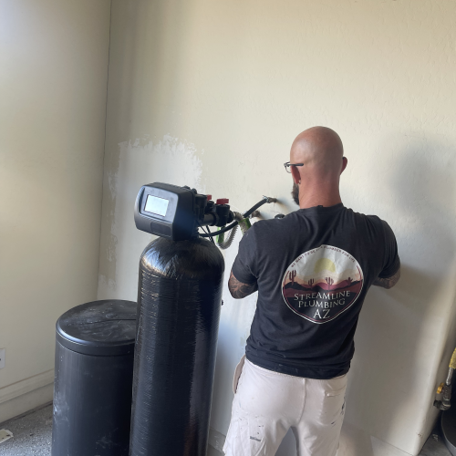 A bald man in a "Streamline Plumbing" t-shirt works on a wall next to a large black water softener system.