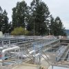 Modern Water Treatment Facility An outdoor view of a water or wastewater treatment plant with multiple open tanks, pipes, and walkways, set against a backdrop of evergreen trees and a body of water.