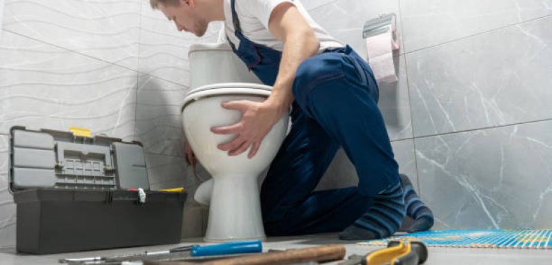 A male plumber in blue overalls and a white t-shirt leans over to fix a toilet in a grey tiled bathroom. A grey toolbox sits open to his left, and various hand tools are scattered on the floor in front of him.
