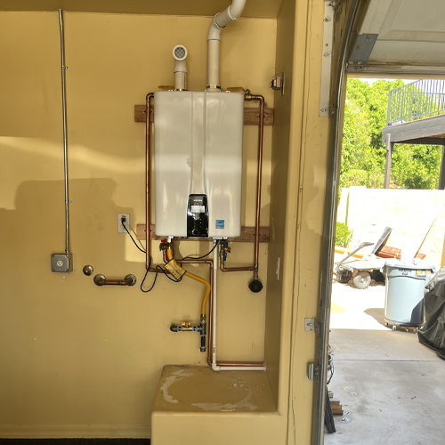 A white tankless water heater is mounted on a yellow wall in a garage, with copper and PVC pipes connected, and an open garage door revealing an outdoor view.