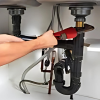 A person's hands using a red wrench to work on the black drain pipes and connections underneath a double kitchen sink.