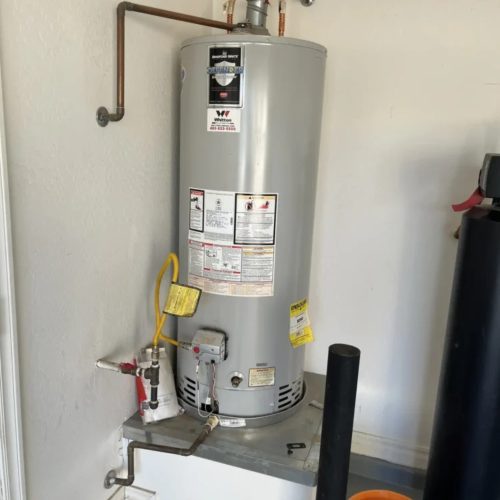 A tall, light gray Bradford White water heater sits on a raised platform, with visible plumbing connections and a nearby water softener or filter unit.