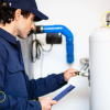 A male technician in a dark uniform and cap inspecting plumbing connections on a white water heater or tank.