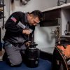 A plumber in a uniform with an American flag patch kneels to work on a garbage disposal unit, with a tool bag open nearby.