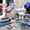 A plumber's hands assemble white plastic drain pipes in a bathroom, with tools and shiny new plumbing parts scattered on the tiled floor.