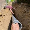 Professional Drainage and Sewer Line Installation A person's hand holds a large grey PVC pipe in a narrow trench while another person works deeper in the trench laying drainage or sewer lines next to a brick wall.
