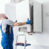 A bald male technician in blue overalls and gloves is installing or servicing a white wall-mounted boiler or water heater, standing on a step ladder.