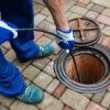 A person in blue work pants and gloves feeds a black hose or snake into an open manhole on a patterned brick patio.