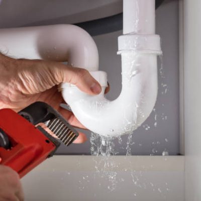 A plumber's hands hold a leaking white PVC pipe under a sink, with water dripping, and a red pipe wrench held nearby.