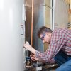 A male technician wearing blue safety glasses and a plaid shirt kneels beside a large water heater, appearing to fix a leak with tools and a rag.