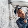 A male technician in a plaid shirt, overalls, and work gloves is using a screwdriver to service or install a white wall-mounted water heater, with tiled walls in the background.